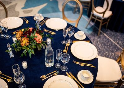 A round table with a dark blue tablecloth is set with elegant dinnerware, glassware, a floral centerpiece, and gold-accented utensils, prepared for a formal gathering at The Cable Center event venue in Denver.