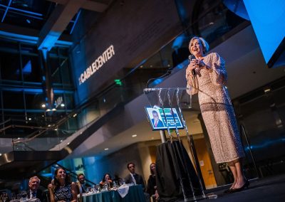 A woman stands at a podium giving a speech in the modern Daniels Great Hall with an audience seated at tables. The background features signage reading "THE CABLE CENTER," and the lighting casts blue hues, highlighting this unique event venue in Denver.