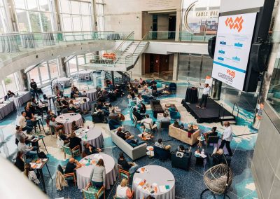 A corporate conference taking place in the elegant, spacious Cable Center event venue with round tables and chairs. Attendees are seated, listening to a speaker at the stage while a large screen displays presentation slides.