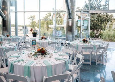 The Cable Center, a unique, spacious, light-filled, and modern Denver event venue, is set up for an elegant occasion with round tables covered in white and turquoise linens, floral centerpieces, and white chairs arranged neatly in the Daniels Great Hall. Large windows reveal a garden outside.