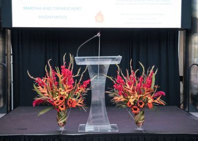 An elegant, clear podium stands on a stage flanked by two large floral arrangements with red and orange flowers. A projector screen in the background displays sponsor names at The Cable Center, a unique Denver event venue.