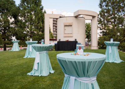 Outdoor event setup with tall round tables covered in light green tablecloths, topped with small decorations, creating an elegant ambiance. Trees and a modern concrete structure are in the background at The Cable Center, a unique Denver event venue.