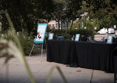 Elegant outdoor event setup at The Cable Center with a bar table draped in black tablecloths. A teal sign on an easel stands nearby. Lush greenery and buildings can be seen in the background, creating a cozy and elegant atmosphere perfect for any Denver event.