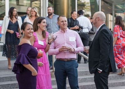 A group of people in formal attire are socializing outdoors, holding drinks and talking. There are stairs and large glass windows of The Cable Center in the background.