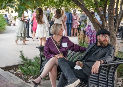 Two people sit on a bench at an outdoor event, both holding drinks. The woman wears a purple dress and the man is in a black suit and cap. A group of people is socializing in the background during this event at The Cable Center in Denver.