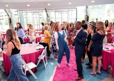 A man in a suit speaks into a microphone while women, some standing and some seated at round tables with pink tablecloths, listen, laugh and applaud at an elegant indoor event held at The Cable Center in Denver.