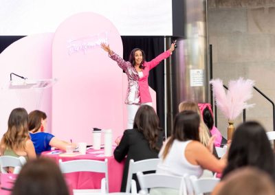A woman stands on a stage decorated with pink elements, raising her arms enthusiastically in front of an audience seated at round tables at The Cable Center - an elegant Denver event venue.
