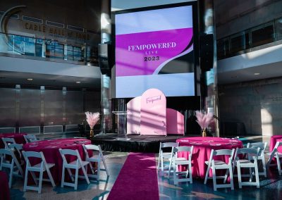 A stage with a large screen displaying "Fempowered Live 2023" graces The Cable Center event venue. The setting features round tables covered with pink tablecloths and white chairs arranged around them, creating a sophisticated atmosphere perfect for an unforgettable Denver event.