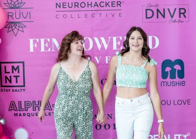 Two women walk hand in hand in front of a pink backdrop with various brand logos at an elegant event. One woman is wearing a green patterned outfit, and the other is in a green crop top and white ripped jeans.