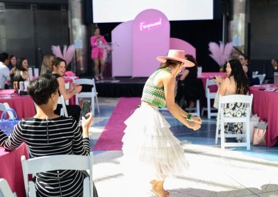 A woman in a white ruffled skirt, green vest, and pink hat dances among round tables draped in pink cloths, while guests seated around clap and take photos of the unique performance.