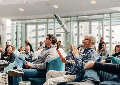 A group of people seated indoors. A man in a flannel shirt speaks into a microphone, holding a book, and others listening attentively in the background.