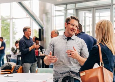 People engaged in conversation at a corporate event in an elegant, modern indoor setting with large windows. A man in a gray shirt gestures while talking to a woman with blonde hair and a brown bag, at The Cable Center event venue in Denver.