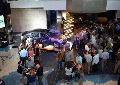 A crowd gathers around a Lexus car display in The Cable Center event venue showroom, engaging in conversations and browsing informational booths. The car is showcased with open doors and illuminated signage, adding to the atmosphere.