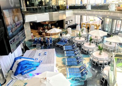 The Cable Center event venue indoor atrium with an pool slide, beach chairs, and round tables with umbrellas. A large screen displays information. Several people are seated, and a food kiosk is in the background.