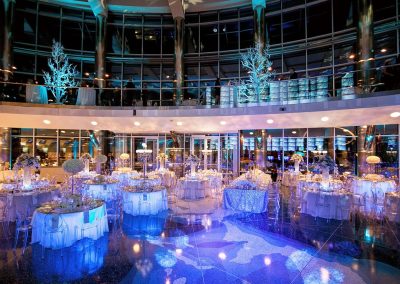 The Cable Center The Cable Center Denver event venue elegantly decorated with multiple round tables set for a banquet in The Daniels Great Hall, featuring high ceilings, large windows, and blue-toned lighting, creating a unique and grand atmosphere for your corporate event.