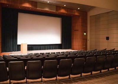The elegant Malone Theater at The Cable Center event venue, featuring rows of black chairs facing a large screen and a wooden podium on stage.