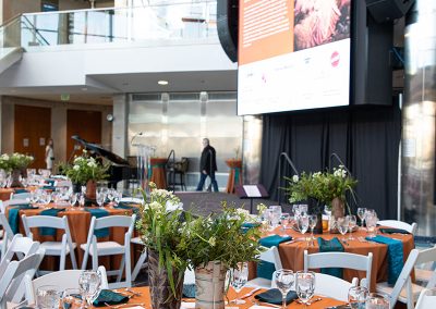 The Cable Center event venue adorned with round tables set with orange tablecloths, green napkins, and glassware, featuring unique floral centerpieces. A large screen displaying event information is in the background.