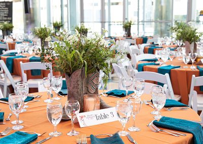 A round banquet table set with an orange tablecloth, teal napkins, glassware, and cutlery awaits guests. The elegant centerpiece features a floral decorations in cowboy boot vases. A table card labeled "Table 3" is placed in the center.