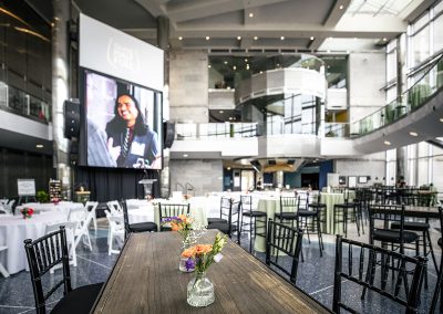 The Cable Center event venue in Denver arranged with round and rectangular tables, black chairs, and a large screen displaying the event title "Glass Half Full" and two people greeting each other. The setting boasts a unique mix of industrial and contemporary architecture, creating an elegant atmosphere perfect for any occasion.