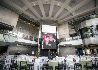 A large, elegant conference room set up for an event with round tables and chairs. A big screen at the front displays an image of two men. The Cable Center’s unique space, boasting high ceilings and modern architecture, makes it a premier Denver event venue.