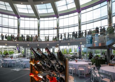 Interior of The Cable Center, a modern circular building with large glass windows. There are rows of wine bottles in the foreground and people socializing on an upper level mezzanine in the background.