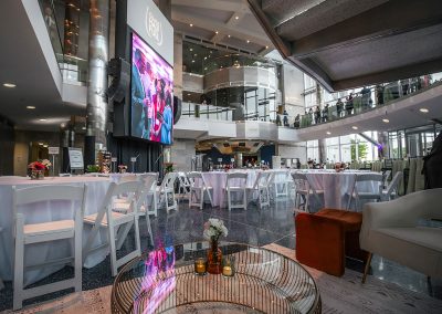 Large atrium with round tables, white chairs, a large screen, with a glass-walled mezzanine above. The room is elegantly set up for a formal gathering, with floral centerpieces and a few scattered lounge chairs.