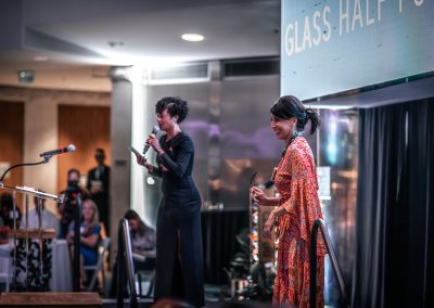 Two women stand on a stage, one speaking into a microphone addressing an audience.