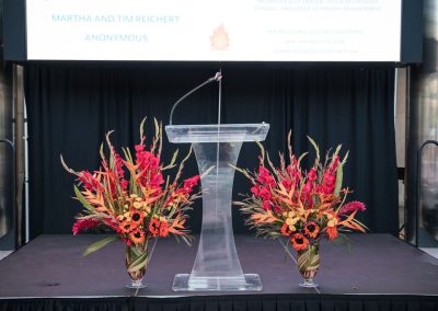 An elegant, clear podium stands on a stage flanked by two large floral arrangements with red and orange flowers. A projector screen in the background displays sponsor names at The Cable Center, a unique Denver event venue.