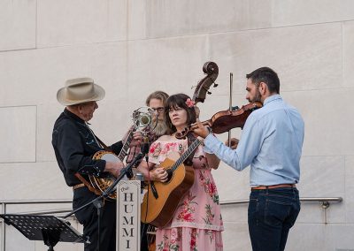 A unique group of musicians performs outdoors. A woman in a floral dress elegantly plays the guitar accompanied by three men playing a banjo, violin and stand up bass at The Cable Center event venue in Denver. A sign in front of the musicians reads “The Humminbirds.”