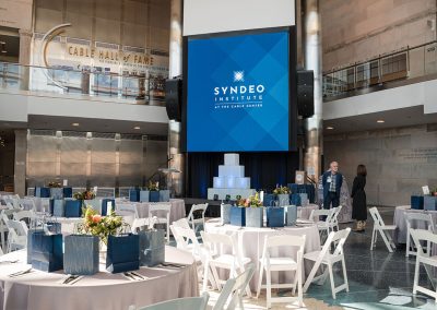 An elegant conference room set with round tables and white chairs, featuring a large screen displaying "Syndeo Institute at the Cable Center." This unique Denver event venue is decorated with flower arrangements and gift bags.