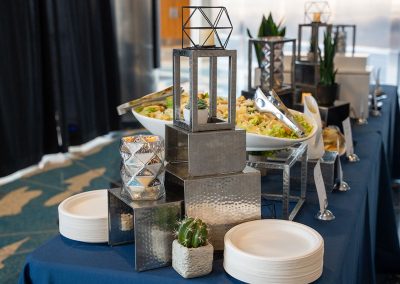 A buffet table with salad in a large white bowl, plates, and metal decor, featuring elegant geometric candle holders and a potted cactus.