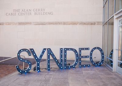 Large illuminated letters spell "SYNDEO" outside the Alan Gerry Cable Center Building.