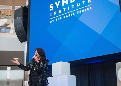 A woman speaks on stage in front of a large blue screen displaying the "Syndeo Institute at The Cable Center" logo. White cube decorations are elegantly stacked near the podium, enhancing the unique ambiance of premier Denver event venue - The Cable Center.