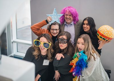 Six people, some wearing colorful costumes and props, gather in front of a photo booth at The Cable Center event venue, looking and posing towards the camera with playful expressions.