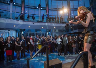 A woman plays the violin on a stage at an elegant indoor event, while an audience stands and watches. The Cable Center event venue in Denver boasts a modern design with glass walls and an upper mezzanine.