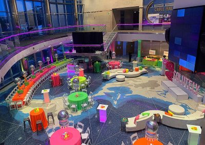 A spacious, brightly decorated event space with colorful tables, chairs, and disco balls. A large map design is visible on the floor, and a sign reading "SOLUTELY" is displayed on a stage. The Cable Center event venue in Denver offers an unforgettable ambiance for any occasion.
