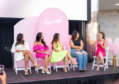 Five women sit on stage, engaging in an elegant author panel discussion. There is a pink backdrop and neon sign that reads “Fempowered”. The woman on the far right is speaking into a microphone, while the others listen attentively.