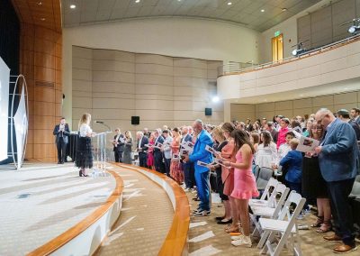 A group of people stands and reads from pamphlets during an indoor event in an auditorium with white, wooden, and carpeted décor. A speaker stands on stage at the podium.