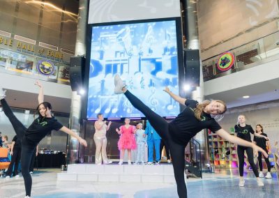 Two dancers perform high kicks in the foreground, showcasing their unique talent. A band plays on stage, with a large screen displaying musical notes behind them. The Cable Centert event venue in Denver features an elegant, tall, modern interior.