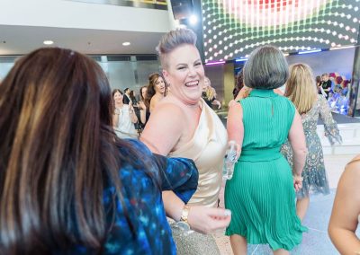 A woman in a cream dress smiles and looks back while locking arms with another woman. Others are also dancing around them. The elegant setting at The Cable Center event venue features a lively indoor setting with a large video screen backdrop.