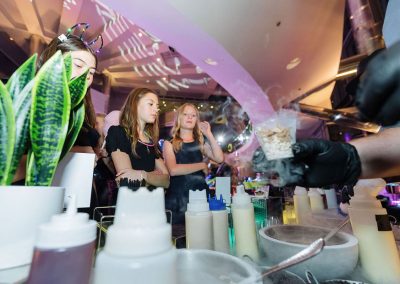 Three girls watch in awe as a person wearing black gloves performs a science experiment with chemicals and dry ice at an elegant, colorful event setup in the unique Cable Center event venue. Various laboratory equipment and plants add to the intriguing display.