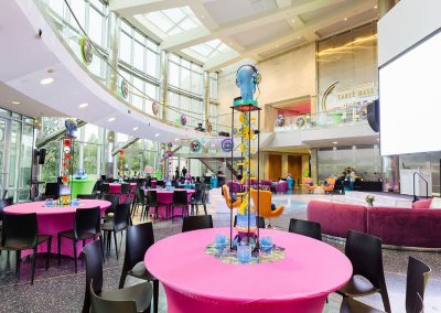 A brightly lit room with high ceilings and large windows, set up for an elegant event with pink tablecloths, black chairs, and colorful decorations. Large screen and a stage are visible in the background of The Cable Center event venue in Denver.