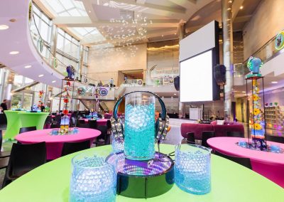 The Cable Center featuring pink and green tables, a large central display with elegant blue decorative elements and headphones, and a large projection screen in the background.