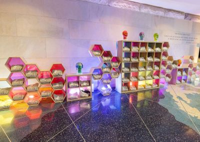 A display wall with hexagonal shelves and cubicles housing goodie bags, elegantly backlit with colorful lights. Five mannequin heads are on top of the cubicles. The dark, glossy mosaic tile floor pattern adds a unique touch, perfect for any stylish Denver event.