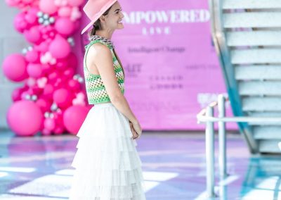 A woman in a pink hat, green and white top, and tiered white skirt walks near a staircase at The Cable Center event venue in Denver. Pink balloons and an "Fempowered" sign appear in the background.