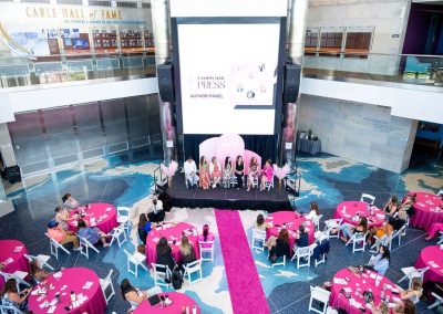 An elegant indoor event is unfolding at The Cable Center event venue in Denver, featuring many round tables draped in pink cloths and an engaging author panel on stage. Attendees are seated around the tables, attentively watching the speakers.