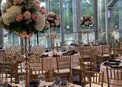 Elegant banquet hall setting at The Cable Center event venue in Denver. There are round tables covered in dark pink tablecloths, set with gold plates and black napkins. Tall floral centerpieces feature white and pink flowers. Large floor-to-ceiling windows surround the room..