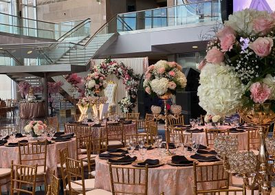 The Cable Center event venue featuring a decorated space with floral centerpieces, pink tablecloths, gold chairs, and elegant lighting. A floral arch and a throne chair stand gracefully near a glass staircase.