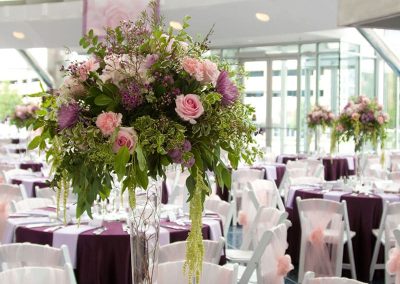 An elegant banquet hall setting at The Cable Center event venue in Denver. There are tables covered in purple tablecloths, set with silverware and menus. Each table features a tall floral centerpiece with pink and purple flowers, while white chairs are draped in pink covers.