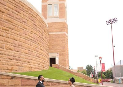 A wedding couple in formal attire walk toward each other near a stone building with a tall tower. The woman wears an intricate gown and veil, and the man is in a black suit.